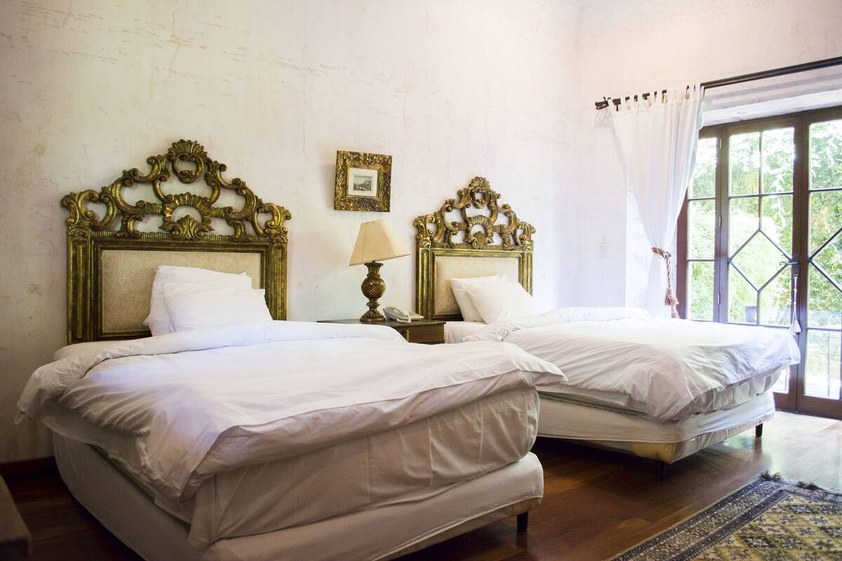 Habitación con dos camas, cada una con cabeceros ornamentados, una mesita de noche con lámpara y una ventana con cortinas.
