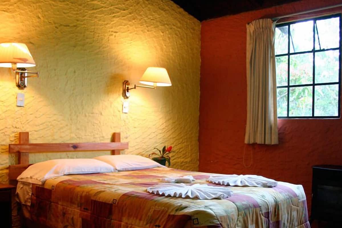 Una habitación acogedora con una cama, dos lámparas de pared, un ventanal con cortinas y una planta pequeña sobre una mesita de noche.