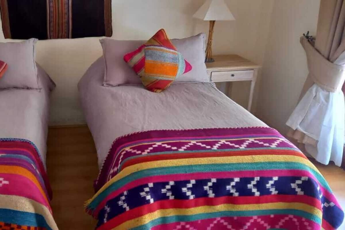 Ein Hotelzimmer mit zwei Betten, jedes mit einer farbenfrohen Decke und einem Kissen. Es gibt Nachttische mit Lampen sowie einen kleinen Tisch mit einer Lampe.