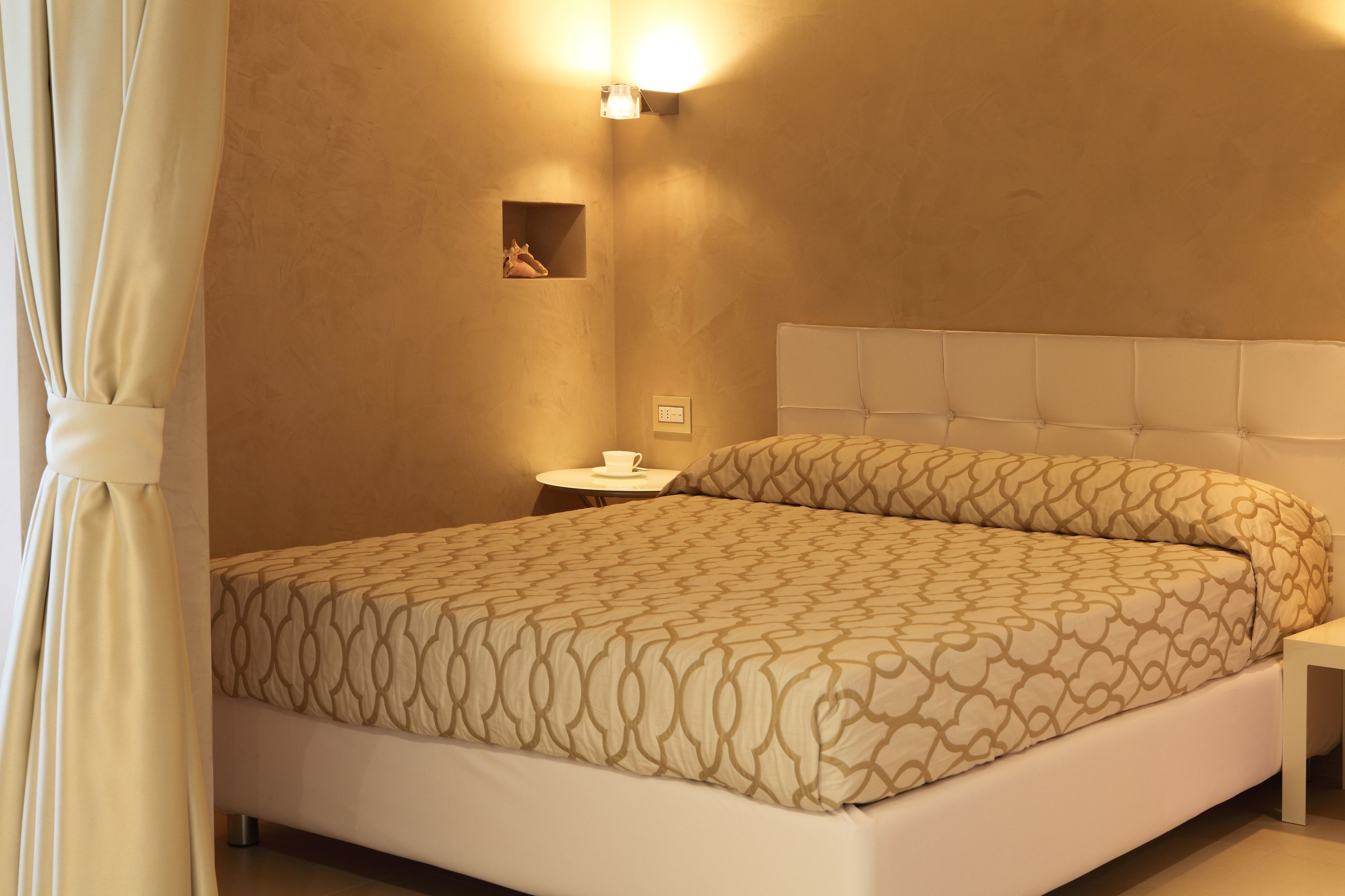 Un lit bien fait, recouvert d’une couvre-lit à motifs, une table de chevet avec une tasse, et un luminaire fixé au mur.