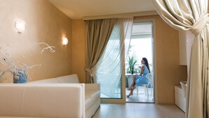 A modern hotel room with a white sofa, a dining table, and a view through a glass door. - Residence Spa Mirabel (Grado)