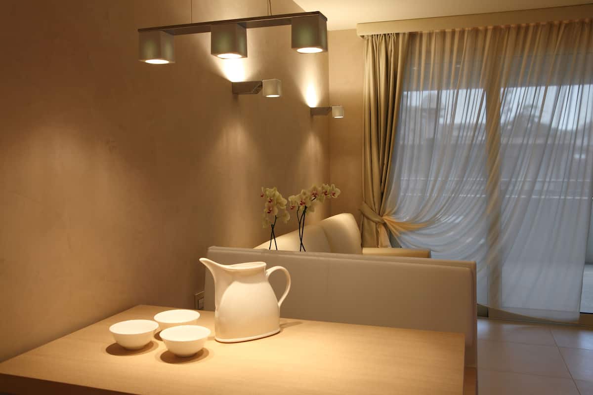 Une pièce moderne avec une table en bois, un pichet blanc et trois bols. On y trouve des lampes fixées au mur et une fenêtre avec des rideaux.