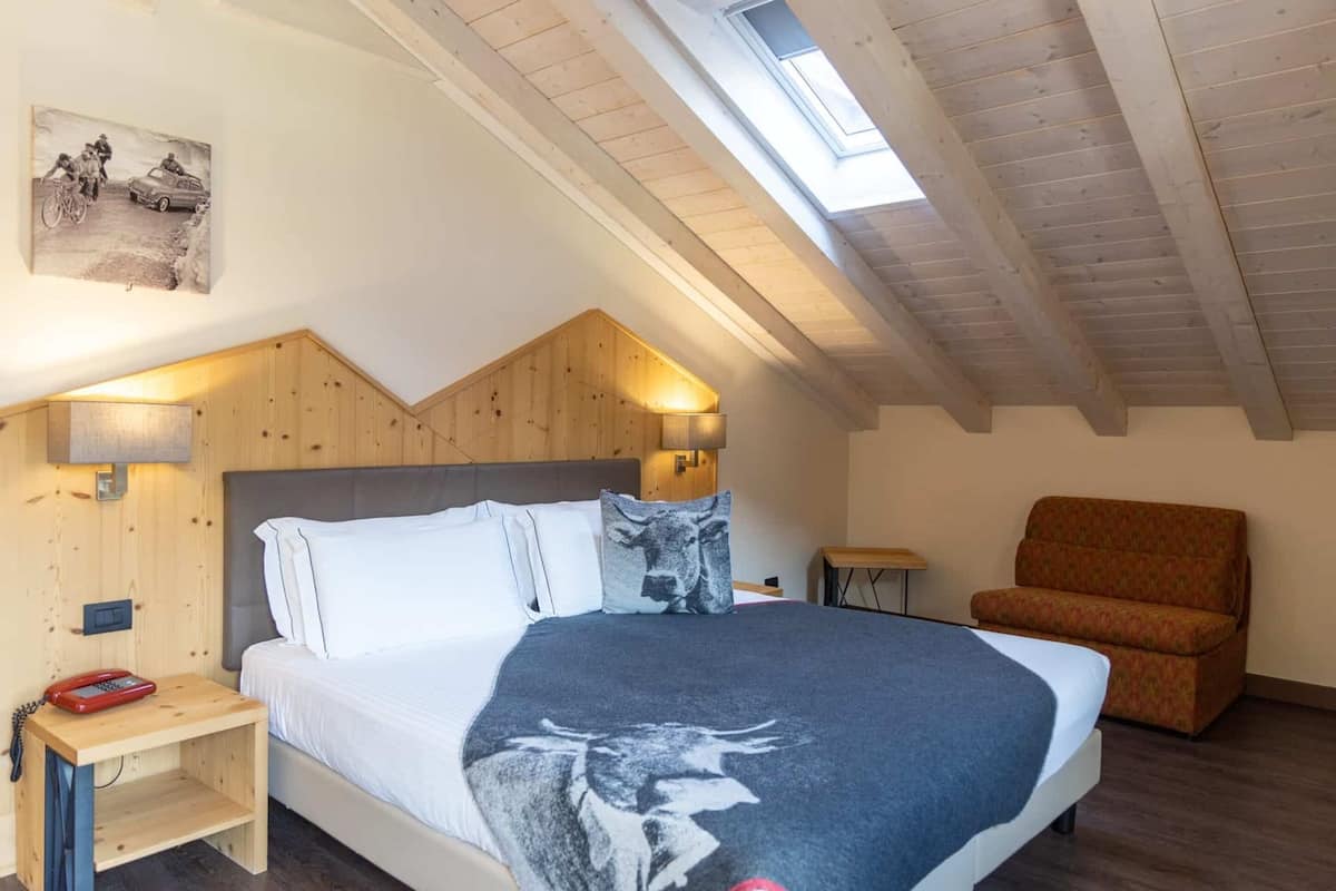 A bedroom with a wooden ceiling, a bed with a gray blanket, a wooden nightstand with a red phone, a small table, and a brown chair.
