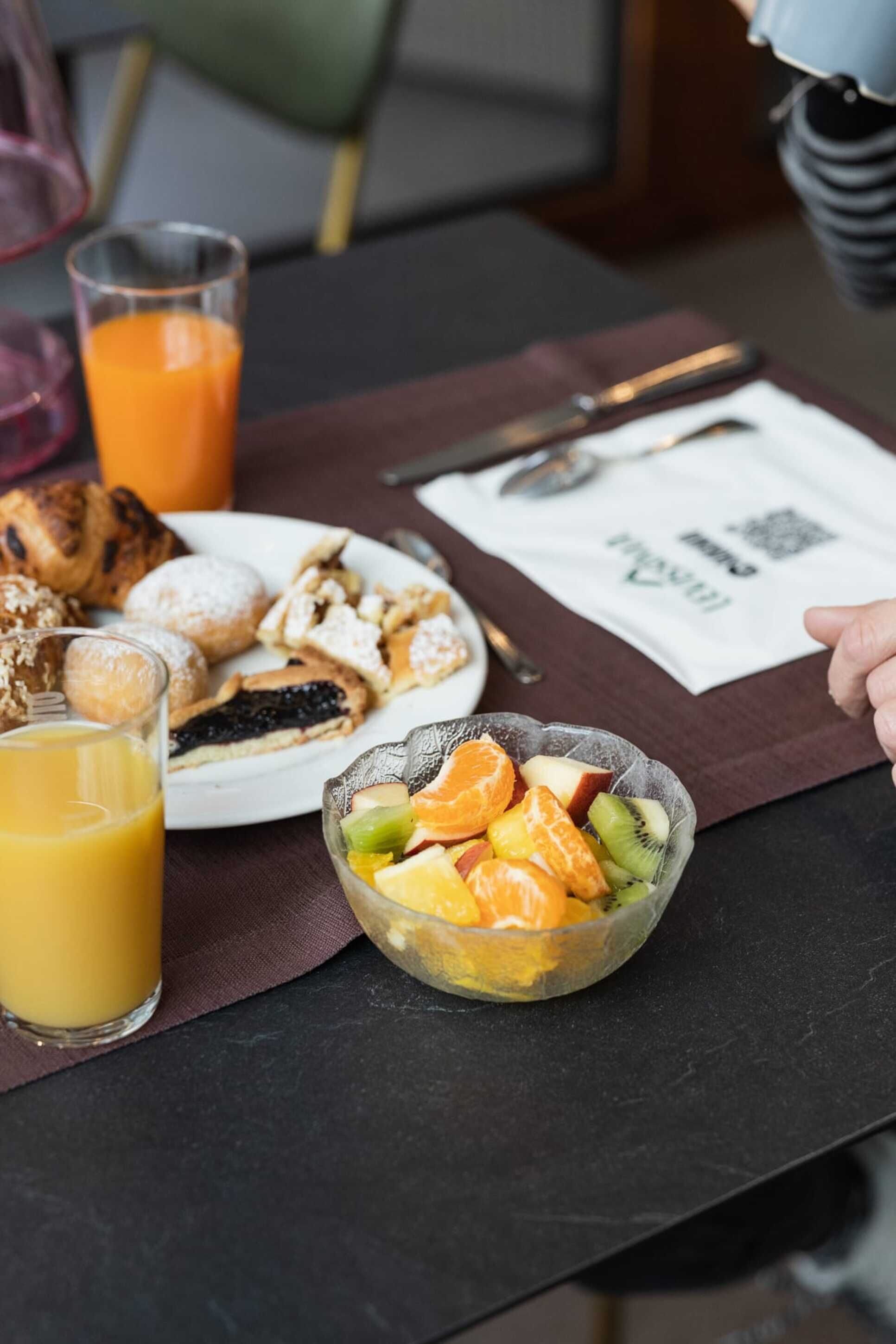 Ett frukostbord med en tallrik bakverk, en skål med frukt, ett glas apelsinjuice och en servett med en QR-kod.