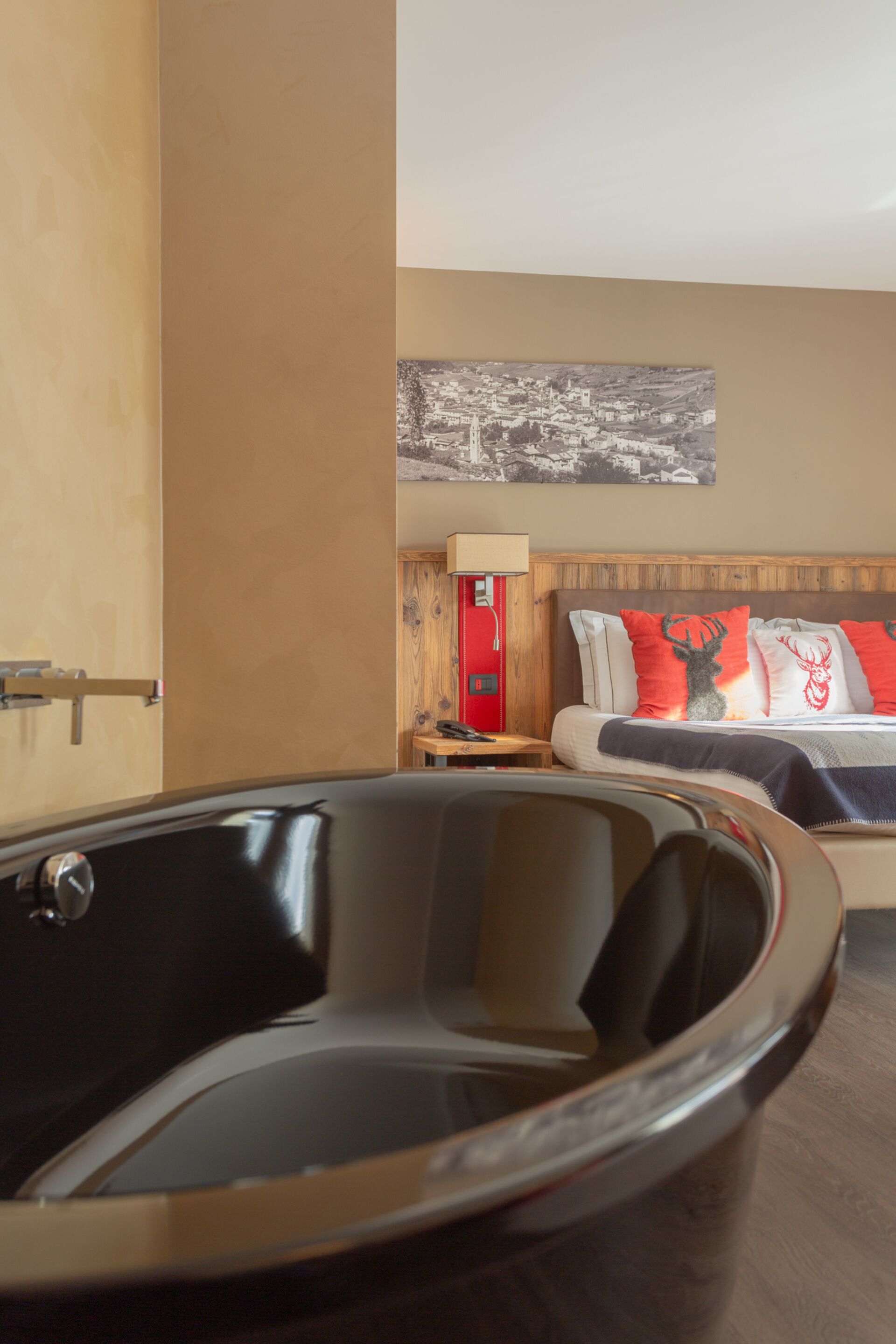 Une chambre d’hôtel moderne avec une grande baignoire noire, une tête de lit en bois agrémentée de coussins rouges et blancs, une petite table de chevet avec une lampe, et un tableau représentant un paysage urbain accroché au mur.