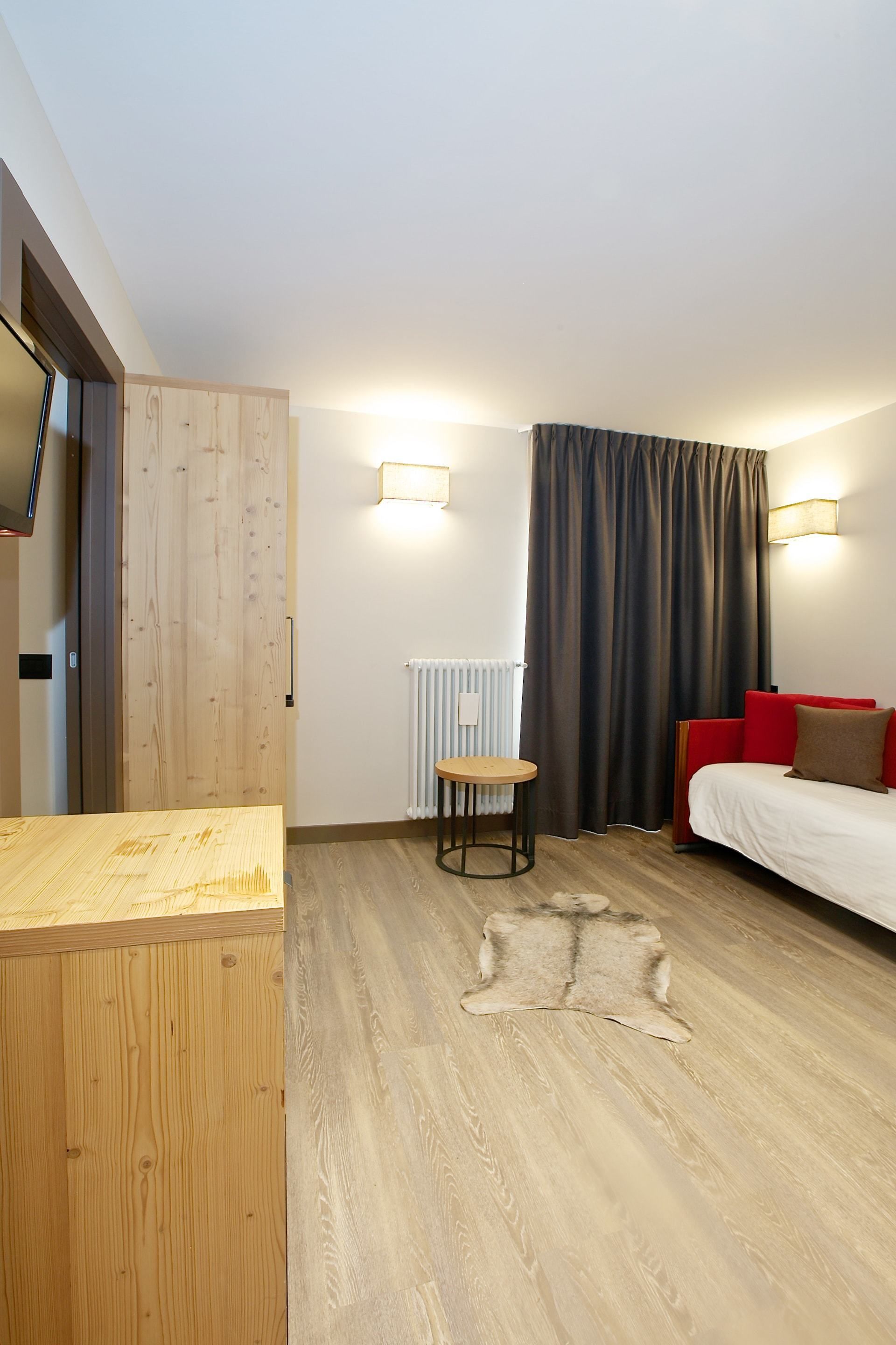 Una habitación moderna con un escritorio de madera, una mesita redonda pequeña, una cama con almohadas rojas y marrones, un ventanal con cortinas y un radiador.