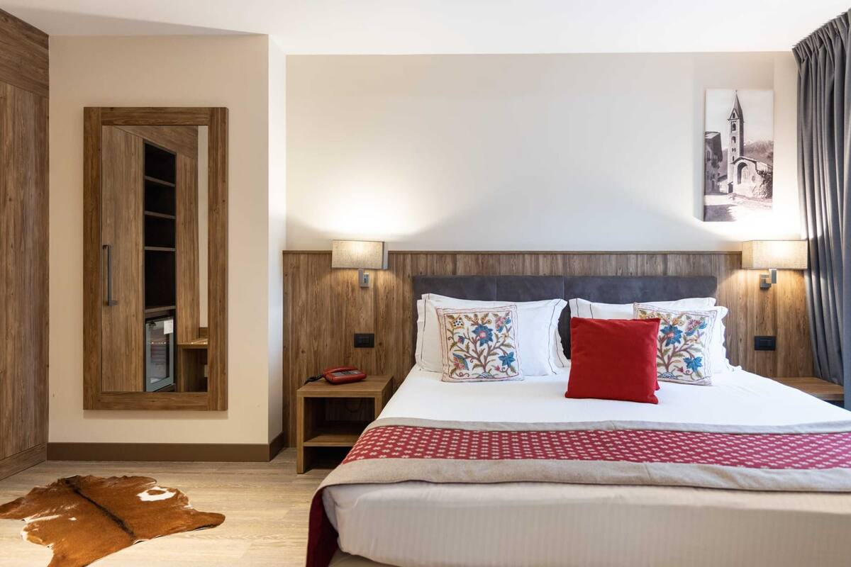 A bedroom with a wooden headboard, a large bed with patterned pillows, bedside lamps, and a mounted picture on the wall.