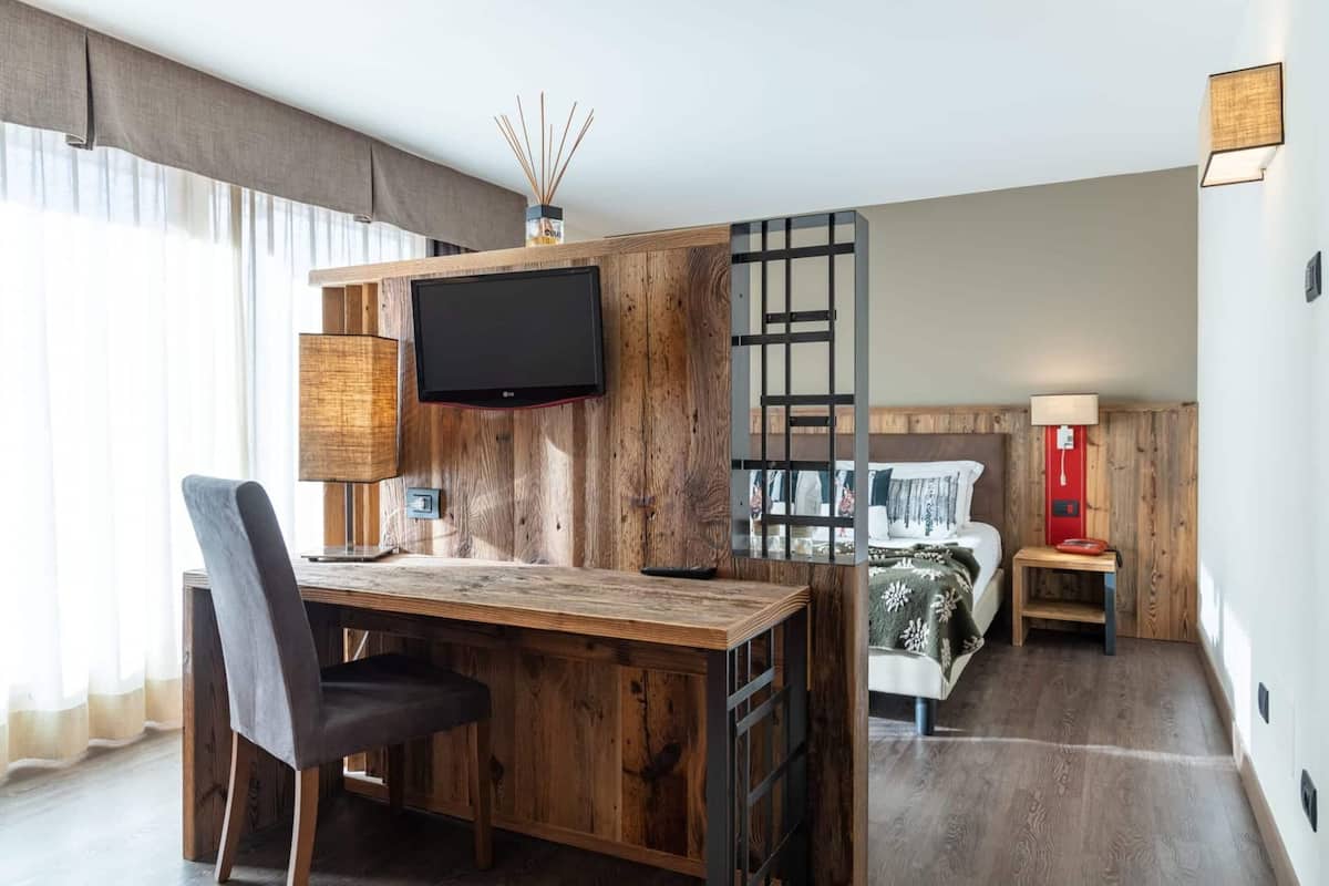 A room with a wooden desk, a chair, a TV mounted on the wall, a bed with a red bedside table, and a nightstand.