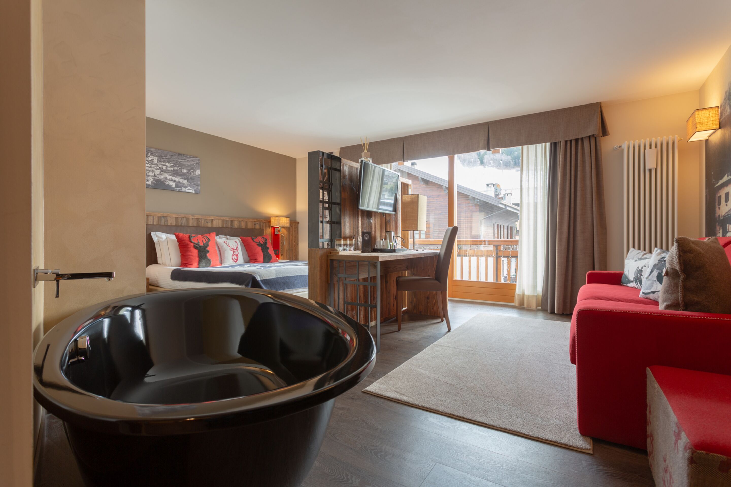 Una habitación de hotel moderna con una bañera negra grande, un sofá rojo, un escritorio de madera y un balcón con vistas a la ciudad.