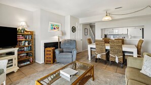 A living room with a television, bookshelf, fireplace, dining area with wicker chairs, and a kitchen in the background. - Endless Ocean Views in S Fletcher Duplex by RedAwning (Fernandina Beach)