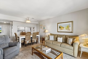 A living room with a sofa, a glass coffee table, a dining area with chairs, a kitchen in the background, and a framed picture on the wall.