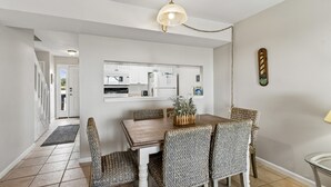 A dining area with a wooden table and wicker chairs, a kitchen in the background, and a ceiling light fixture. - Relaxed Atmosphere in S Fletcher Duplex by RedAwning (Fernandina Beach)