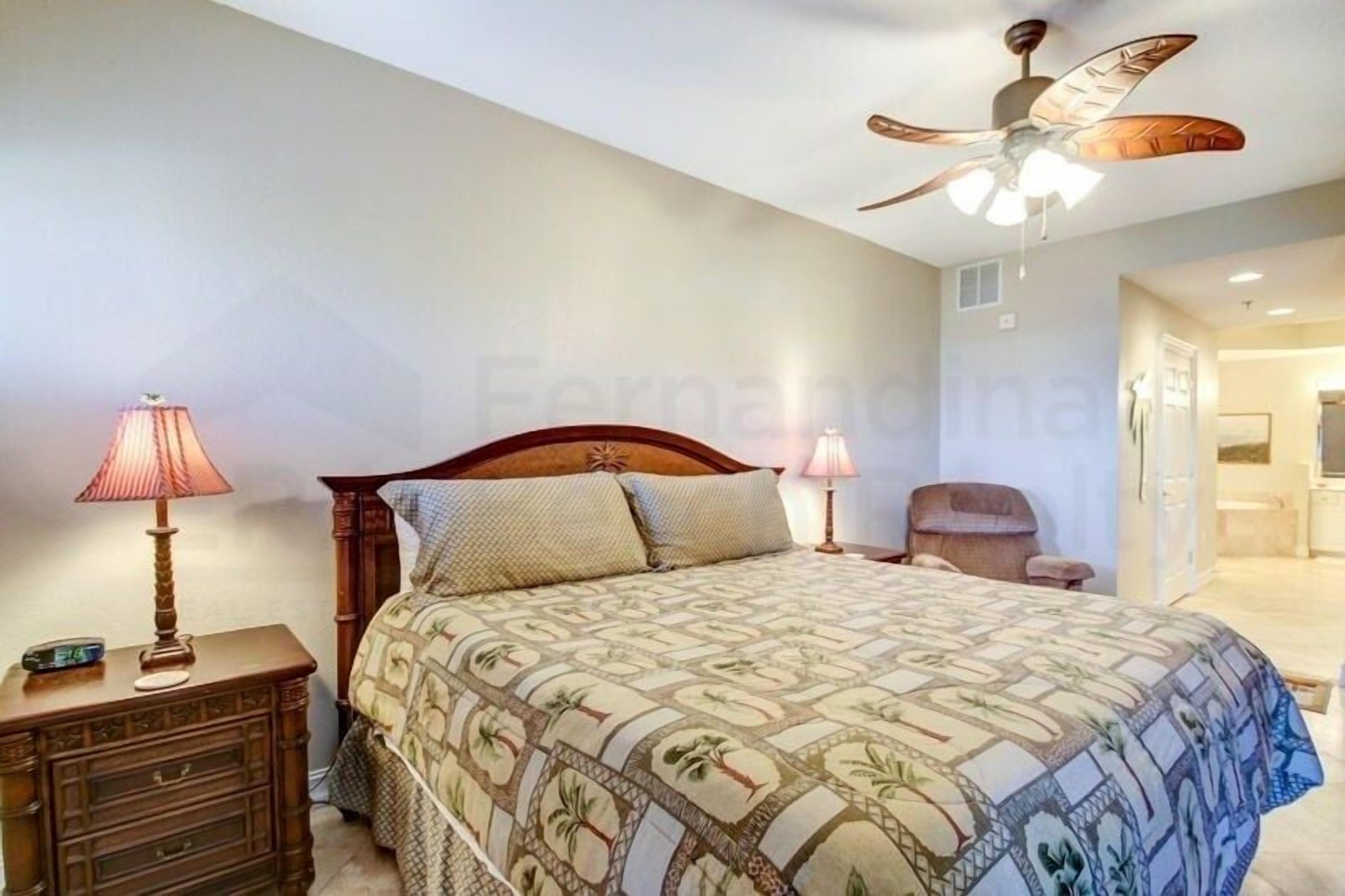 A bedroom with a bed, two bedside lamps, a wooden nightstand, a ceiling fan, and a chair.