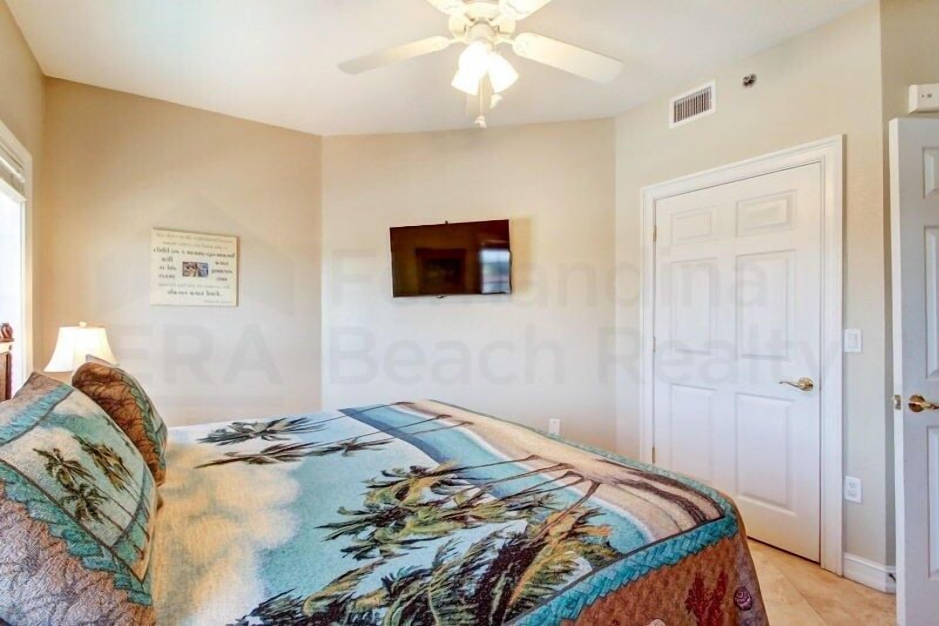 A bedroom with a bed, a ceiling fan, a door, and a wall-mounted TV.