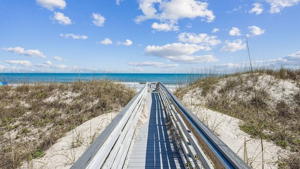 Un muelle de madera que conduce a una playa con dunas de arena y un cielo azul despejado.