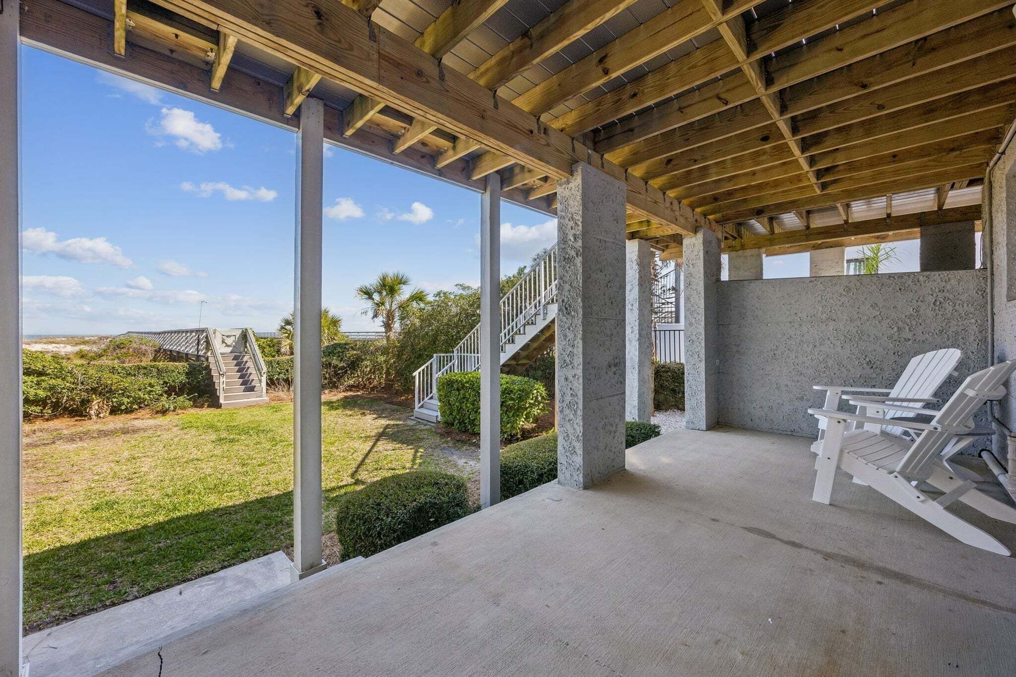 Un espacio al aire libre techado con piso de concreto, una silla Adirondack blanca y una escalera que conduce a un nivel superior.