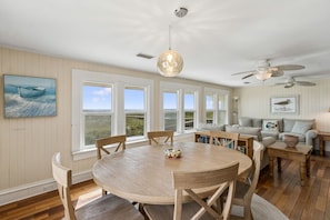 Amplo quarto com uma mesa de jantar redonda de madeira, ventoinha no teto e grandes janelas com vista para o exterior.