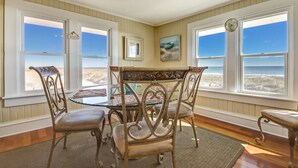Un comedor con una mesa y sillas, ventanas amplias con vista a la playa y un reloj en la pared.