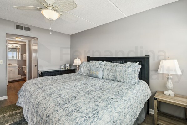 A bedroom with a large bed, a ceiling fan, a nightstand with a lamp, and a door leading to a bathroom. - Amelia South 2 Bedroom Condo by RedAwning (Fernandina Beach)