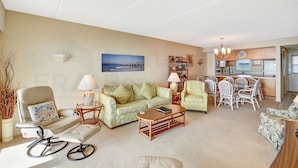 A living room with a sofa, chairs, a coffee table, and a dining area with a table and chairs. There is a kitchen in the background. - Breathtaking Ocean View Amelia South Condo by RedAwning (Fernandina Beach)
