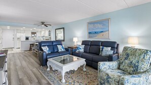 A living room with a sofa, armchairs, a coffee table, and a painting of a beach scene. - Striking Ocean View Condo with Private Walkway to the Beach by RedAwning (Fernandina Beach)
