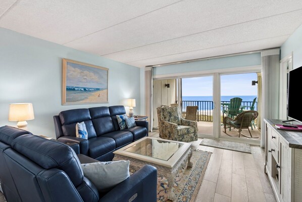 A living room with a sofa, armchairs, a coffee table, and a TV. There is a balcony with outdoor furniture and a view of the ocean.