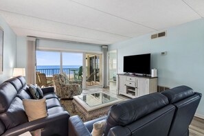 A living room with a sofa, chairs, a coffee table, and a TV. There is a balcony with a view of the ocean. - Striking Ocean View Condo with Private Walkway to the Beach by RedAwning (Fernandina Beach)