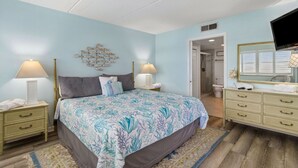 A bedroom with a bed, bedside tables, a dresser, a wall-mounted fish decoration, a television, and a bathroom visible through an open door. - Striking Ocean View Condo with Private Walkway to the Beach by RedAwning (Fernandina Beach)