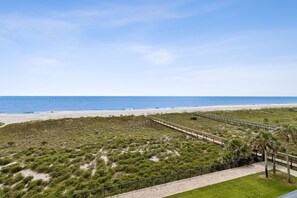 Un paisaje costero con una playa arenosa, un paseo marítimo y una zona cubierta de césped con vegetación escasa.