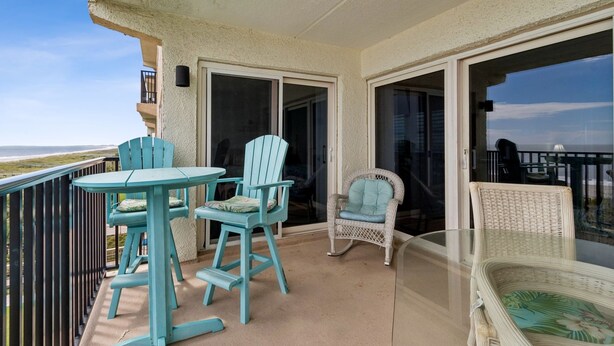 A balcony with a table and chairs, a wicker chair, and a glass table. - Striking Ocean View Condo with Private Walkway to the Beach by RedAwning (Fernandina Beach)