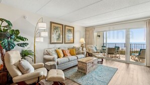 Een ruime woonkamer met een grijze bank, beige fauteuils en een salontafel. Er zijn twee grote ramen met uitzicht op het strand.
