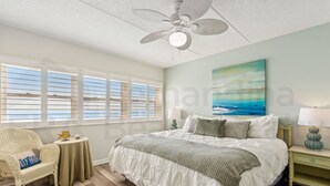 Een slaapkamer met een bed, een stoel, een klein tafeltje, een nachtkastje en een plafondventilator.