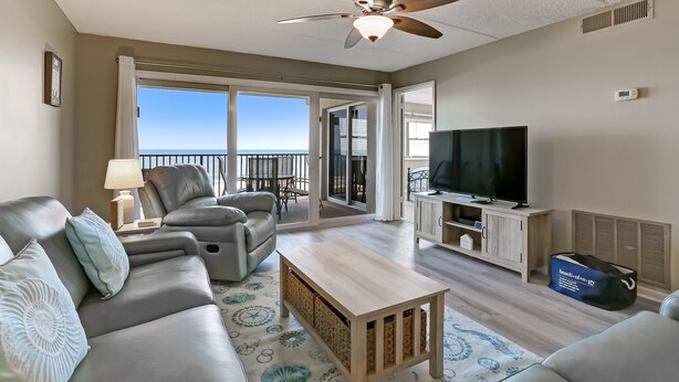 A living room with a sofa, a coffee table, a TV, and a ceiling fan. - Enhanced Unit with Awesome View of Atlantic from Private Balcony by RedAwning (Fernandina Beach)