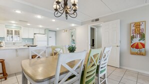 A dining area with a wooden table and white chairs, a chandelier, and a painting on the wall. - Condo with Atlantic View from Private Patio, Short Walk to the Beach by RedAwning (Fernandina Beach)