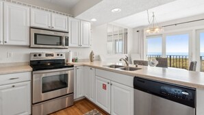 A modern kitchen with white cabinets, stainless steel appliances, a dishwasher, and a view of the ocean through large windows. - Renovated Seaside Condo with Gorgeous View of the Ocean by RedAwning (Fernandina Beach)