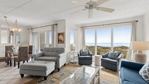 A spacious living room with a large window offering a view of the beach, a ceiling fan, and a variety of seating options including a sofa, armchairs, and ottoman. - Renovated Seaside Condo with Gorgeous View of the Ocean by RedAwning (Fernandina Beach)