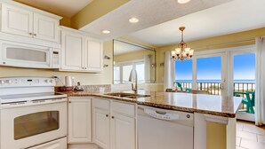 A kitchen with white cabinets, a built-in microwave, and a dishwasher. There is a large window with a view of the beach.