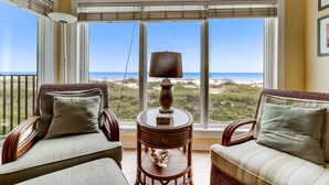 A room with wicker furniture, a round table with a lamp, and large windows with a view of the beach.