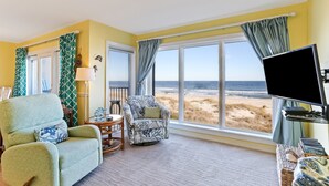 Quarto com uma grande janela com vista para a praia, um ventilador de teto e duas poltronas com almofadas estampadas.