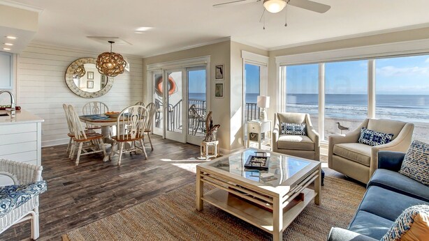 A spacious living room with a large window overlooking the ocean, a ceiling fan, and a dining area with a round table and chairs.