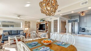 A dining area with a wooden table, white chairs, and a bowl of fruit. There is a ceiling fan, a television, and a kitchen in the background.