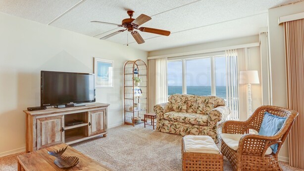 A living room with a television, wicker furniture, a ceiling fan, and a view of the ocean. - Coastal Condo with Ocean Viewing Pleasure by RedAwning (Fernandina Beach)