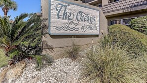 A sign for "The Oceans of Amelia" with a coastal theme, surrounded by desert plants and a building in the background. - Coastal Condo with Ocean Viewing Pleasure by RedAwning (Fernandina Beach)