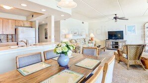 A modern kitchen with wooden cabinets, a refrigerator, and a dining area with a wooden table and chairs. - Coastal Condo with Ocean Viewing Pleasure by RedAwning (Fernandina Beach)