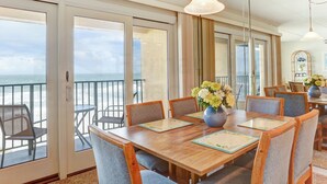 A dining area with a wooden table, chairs, and a view of the ocean through large glass doors. - Coastal Condo with Ocean Viewing Pleasure by RedAwning (Fernandina Beach)