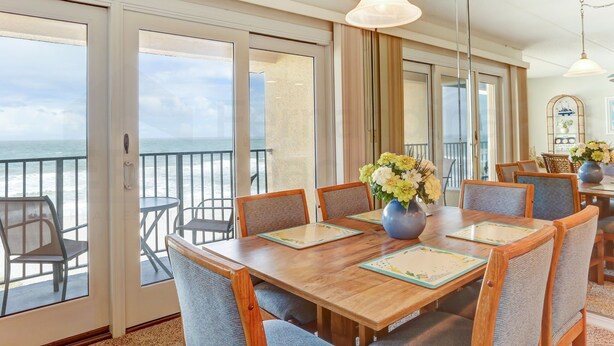A dining area with a wooden table, chairs, and a view of the ocean through large glass doors. - Coastal Condo with Ocean Viewing Pleasure by RedAwning (Fernandina Beach)