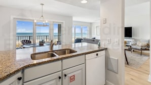 A modern kitchen with a granite countertop, stainless steel sink, and white cabinetry. There is a dining area with a table and chairs, and a living room with a sofa and a television.