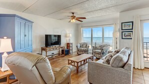 A living room with a TV, seating, a ceiling fan, and a view of the ocean. - Modest Condo with Splendid Sunrise Views by RedAwning (Fernandina Beach)