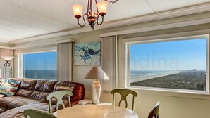 A room with a large window offering a view of the beach, a brown leather sofa, a wooden table with chairs, and a decorative lamp. - Amelia South B7 by RedAwning (Fernandina Beach)