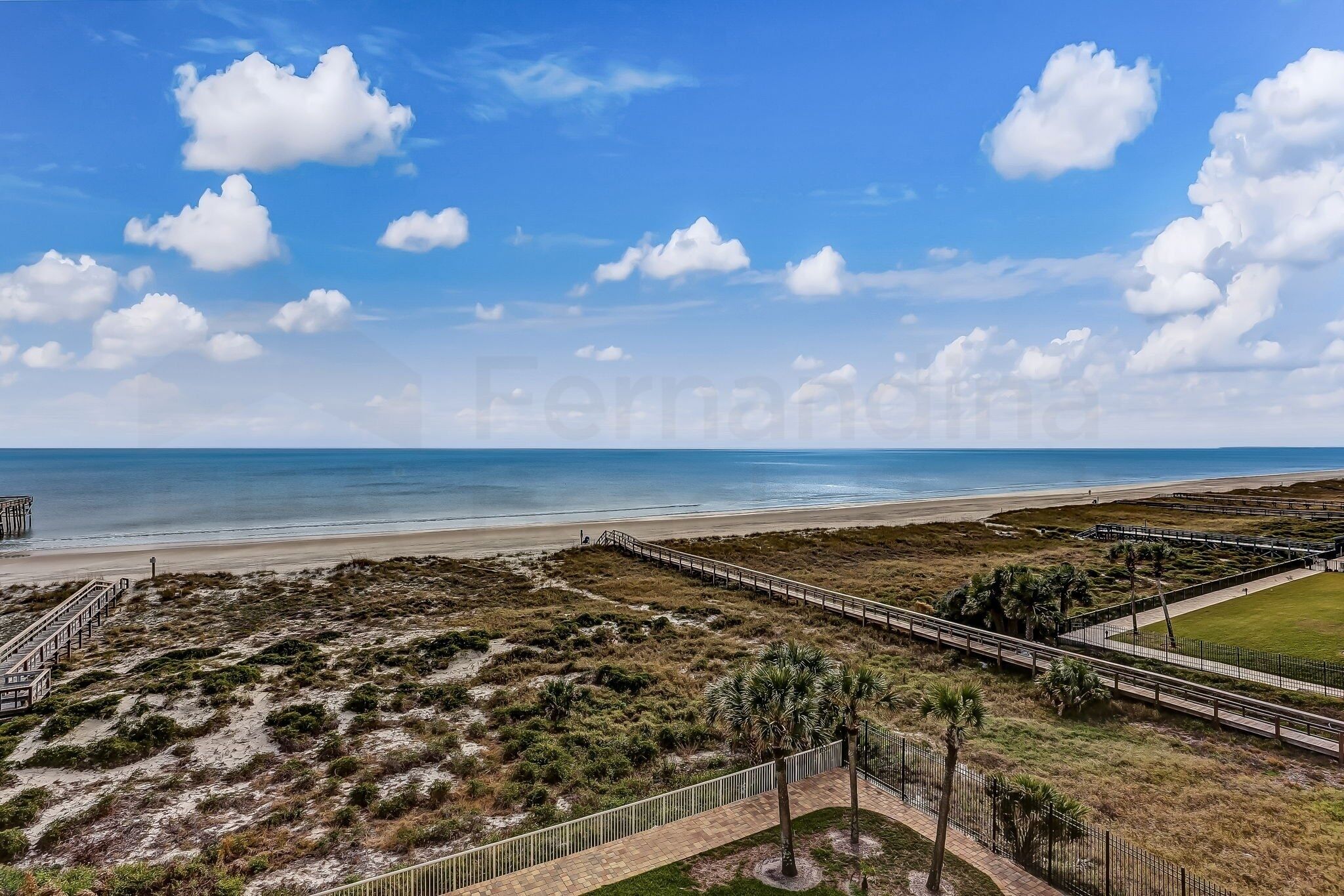 Un paysage côtier comprenant une plage, une promenade en bord de mer et une zone herbeuse parsemée de palmiers.